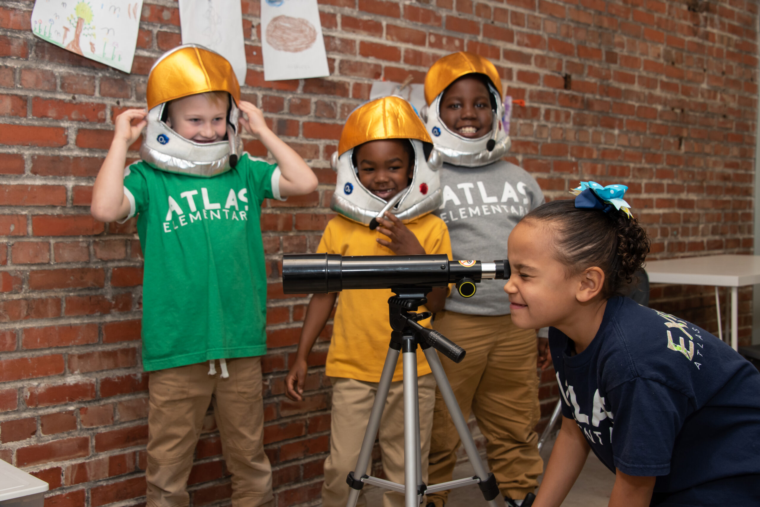 a diverse group of young children, three wearing astronaut helmets and observing a girl looking through a small telescope.